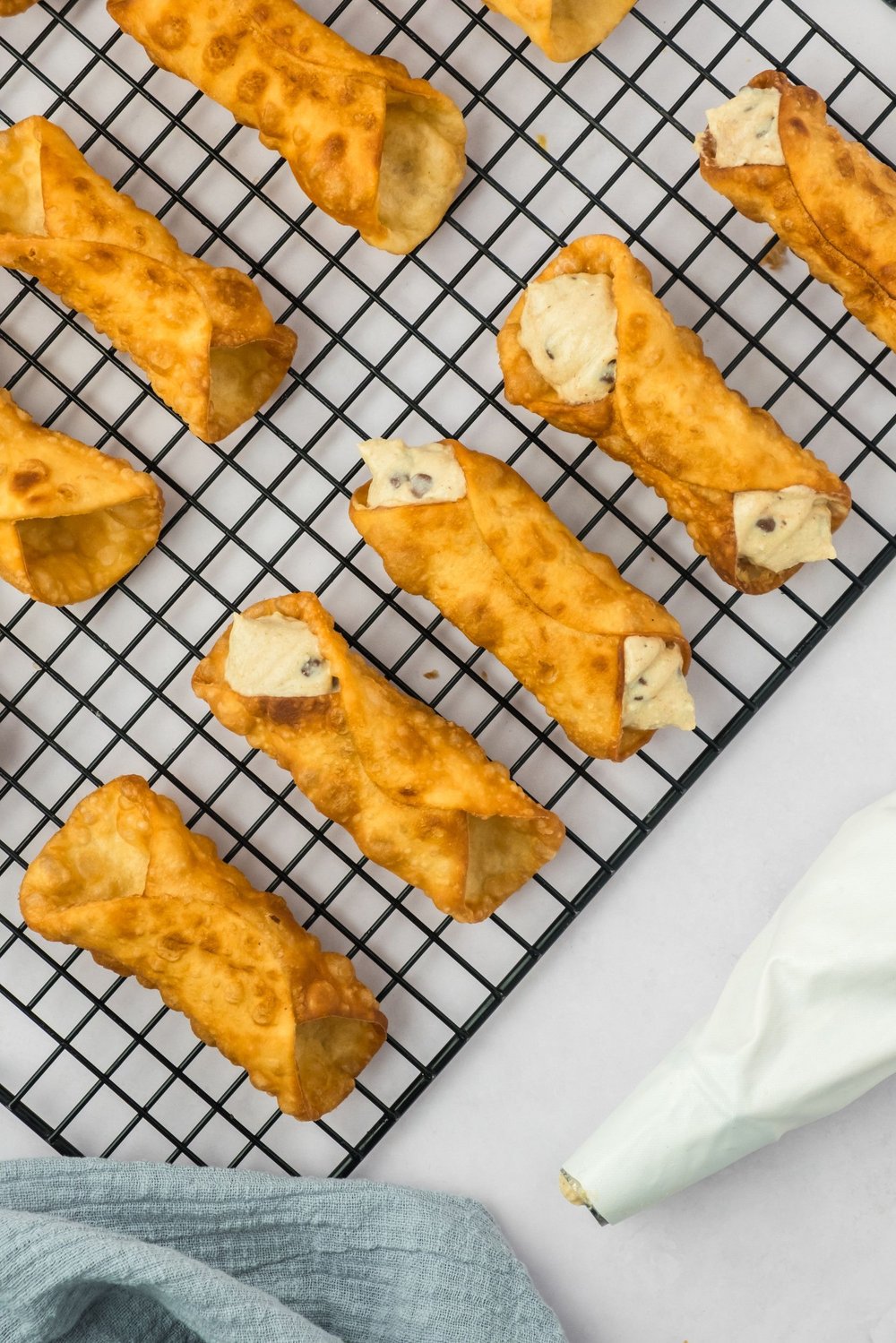 Cannoli shells on a wire rack, some with filling piped in, and a piping bag to the side.