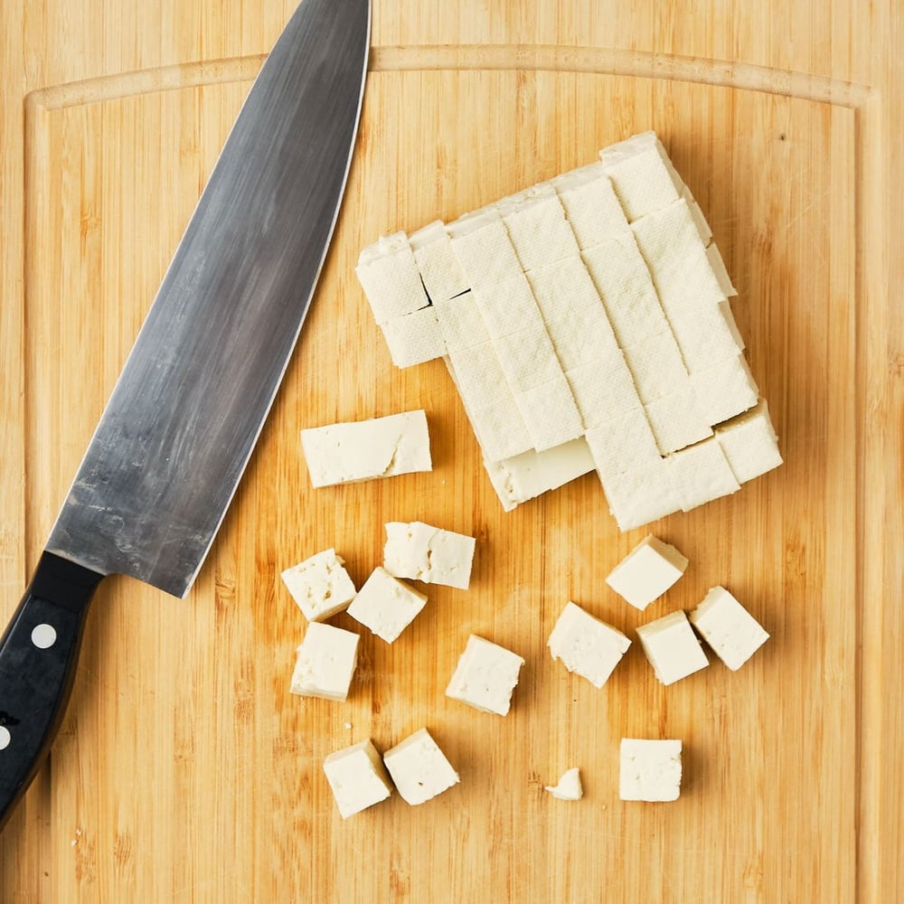 Tofu on a cutting board with a knife, cut into small cubes.