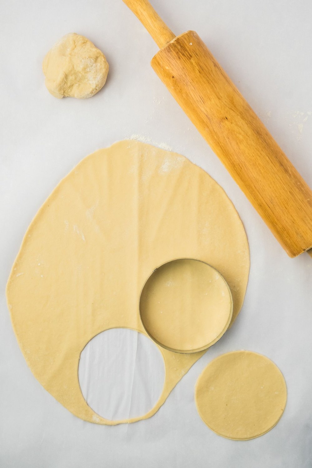 A ball of dough next to rolled out dough, with a rolling pin to the side, and a round cutter on top; one round piece of dough has already been cut.