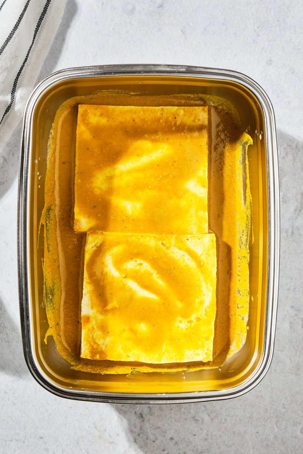Two slices of tofu marinating in a shallow container.