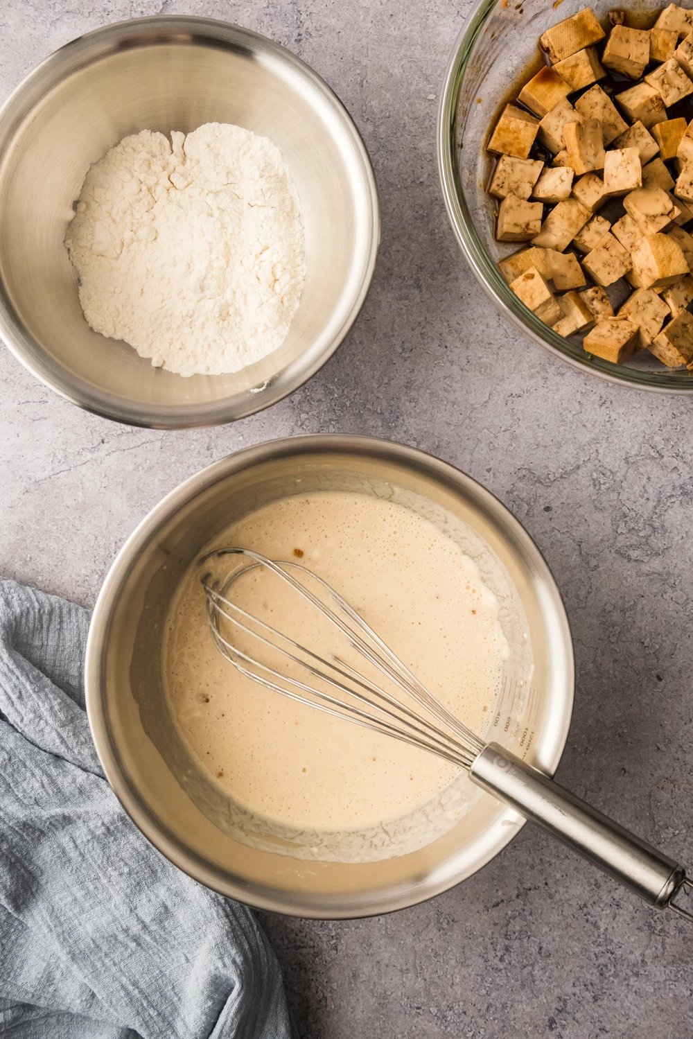 Three bowls on a table: one with mixed tofu batter, one with flour, and one with marinating tofu.
